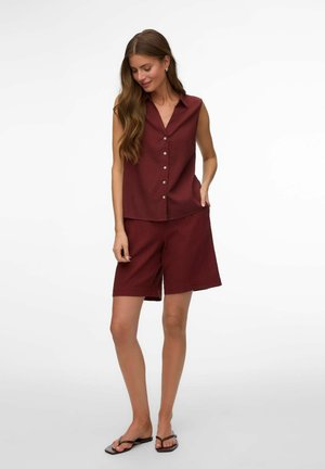 Kvinde iført ærmeløs burgunderrød skjorte med knapper og matchende knælange shorts samt sorte flipflops, stående foran hvid baggrund.