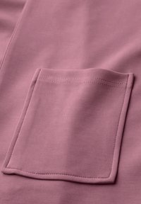 Bolsillo frontal cuadrado cosido sobre una tela suave de color malva, con costura visible y bordes doblados.