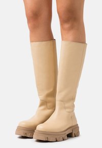 Hnedé kožené čižmy po kolená s hrub�ými podrážkami, nosené na holých nohách, postavené na bielom pozadí.