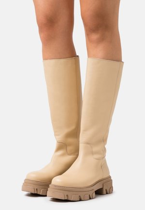 Ceļgala auguma bēšu ādas zābaki ar biezām, raupjām zoleēm, uz kailām kājām, stāvot uz baltas fona.