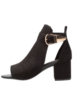 Sort ruskind ankle støvle med åben tå, der har en kraftig blokhæl og en guldlåsdetalje på siden. Glat finish, elegant design.