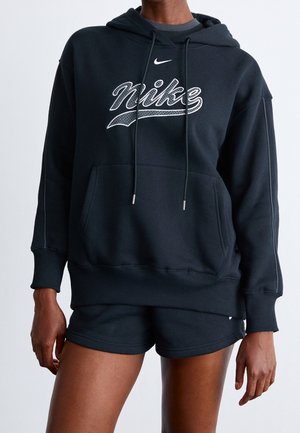 Osoba ubrana w czarną bluzę z kapturem Nike z białym logo oraz czarne spodenki, stojąca na jednolitym, jasnoszarym tle.
