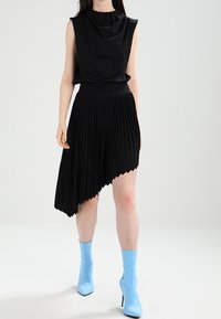 Top negro sin mangas con escote drapeado combinado con una falda negra plisada, que presenta un dobladillo asimétrico. Botines azules de tobillo complementan el look.
