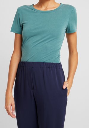 Personne portant un t-shirt ajusté à manches courtes couleur sarcelle, rentré dans un pantalon droit bleu marine à taille élastique, avec la main droite dans la poche, sur fond gris.