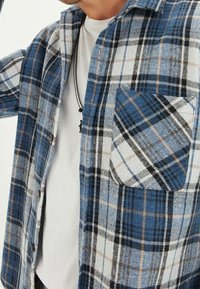 Camicia di flanella con motivo a quadri blu e bianco, con una tasca sul petto e chiusura con bottoni, indossata sopra una t-shirt bianca.