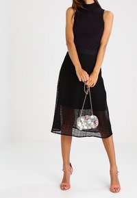 Top negro sin mangas combinado con una falda midi de encaje negra. Presenta un bolso transparente adornado con flores y tacones de tiras en rosa claro.