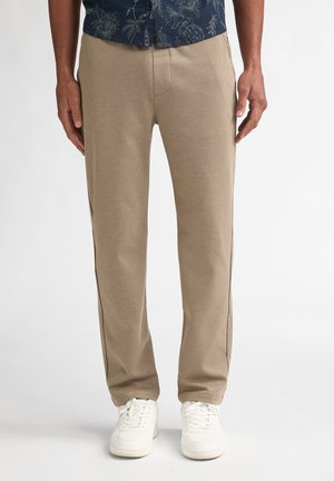 Mann trägt beige gerade geschnittene Hose, weiße Sneaker und ein dunkelblaues, blumiges Hemd, steht vor einem weißen Hintergrund.