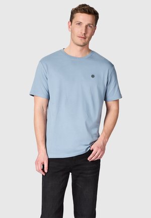 Man draagt een lichtblauw T-shirt met korte mouwen en klein zwart scheepsroerlogo, gecombineerd met een zwarte broek, staand tegen een effen achtergrond.