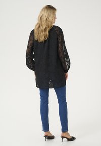 Black sheer blouse with floral pattern, long sleeves, and a loose fit, paired with blue skinny jeans and black heeled mules.