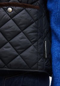 Fekete steppelt szövetkabát barna kordbársony zsebszegéllyel és ezüst patentzárral. Tartalmaz egy fekete márkajelzéses címkét. Kék texturált pulóver fölött viselve.