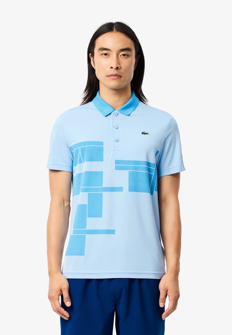 Ljusblå polo med knäppt krage. Har geometriska blå mönster. Tillverkad av slät, andningsbar tyg. Lacoste-logotypen visas.