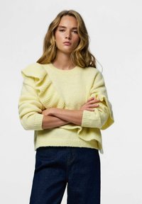 Sweter z krótkim rękawem w jasnym żółtym kolorze z teksturowanym, marszczonym detalem na lewym ramieniu i ściągaczami na rękawach, zestawiony z ciemnoniebieskimi dżinsami.