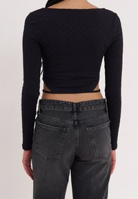 Svart långärmad topp med texturerad tyg, med fyrkantig halsringning och justerbara band i ryggen, ihop med högmidjade grå jeans.