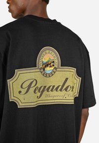 Melnas kokvilnas t-krekls ar vintage stila grafiku aizmugurē, kurā attēlots teksts "Pegador" un kalnu ainava bālgans krāsās.