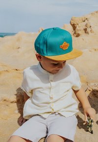 Türkiz snapback sapka sárga karimával, barna logóval; gyermek világos színű pólót és rövidnadrágot visel, játékot tart a kezében.