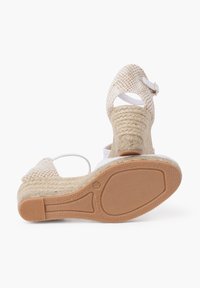 Wedge sandals featuring a woven beige upper, white straps, and a textured natural jute heel. Rubber outsole with patterned grip.
