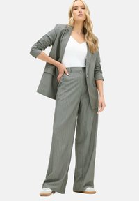 Kvinde iført grå stribet blazer og vide bukser med hvid top og sneakers, stående med den ene hånd på hoften.