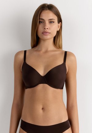 Vrouw met steil lichtbruin haar die een donkerbruine beha met voorgevormde cups en bijpassend laagzittend ondergoed draagt, staand tegen een witte muur.
