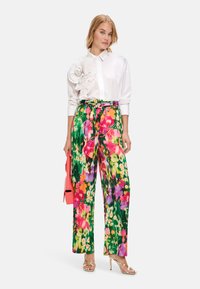 Weißes Hemd mit Knöpfen und einer strukturierten floralen Akzentuierung auf einer Schulter. Bunte, floral bedruckte weite Hose; glänzende silberne Absatzsandalen.