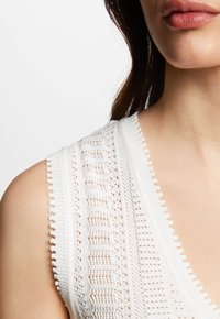 Top in maglia bianca con scollo a V, dettagli intricati di lavorazione aperta e bordi ondulati sulle spalle.