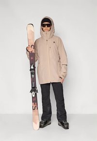 Beige wasserdichte Skijacke mit Frontreißverschluss und Kapuze, kombiniert mit schwarzer Schneehose und Skischuhen. Hält helle Skier in der Hand.