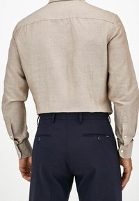 Camicia di lino marrone chiaro con colletto, maniche lunghe e polsini con bottoni, abbinata a pantaloni blu navy con tasche posteriori e dettaglio con bottone.