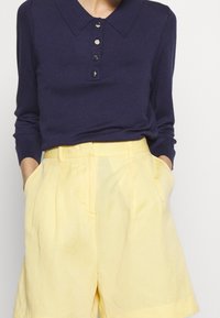 Mörkblå långärmad polo med krage och fem metallknappar, tillsammans med ljusgula högmidjade shorts med sidofickor.