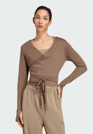 Vrouw draagt een bruin wikkel-lange-mouwen-topje gestrikt in de taille en beige Adidas joggingbroek met een hoge elastische tailleband.