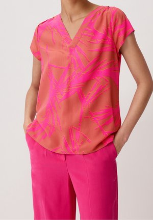 Femme portant un chemisier corail manches courtes avec un motif abstrait rose vif et un pantalon taille haute rose vif, mains dans les poches.