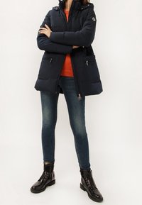 Person med en svart puffjacka över en orange tröja, blå tighta jeans och svarta snörade ankelboots, stående med armarna i kors.
