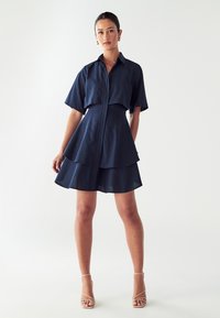 Tamsiai mėlyna suknelė su apykakle, sagčių priekyje ir trumpomis rankovėmis, turinti sluoksniuotą sijoną. Lygi medžiaga su struktūruota siluetu.