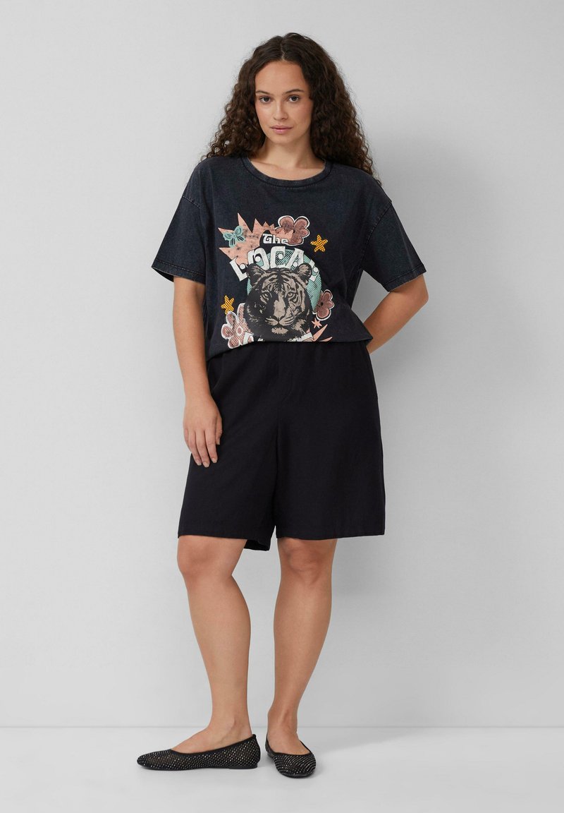 Zwarte katoenen shorts gecombineerd met een losse, donkere T-shirt met grafisch ontwerp van een kleurrijke tijger en bloemaccenten. Zwarte instappers met stippen.
