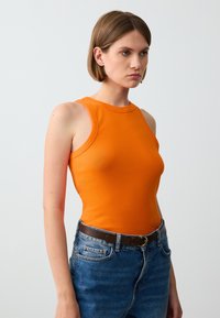 Top aderente arancione con scollatura tonda e ampie aperture per le braccia, abbinato a jeans in denim blu a vita alta e una cintura marrone.