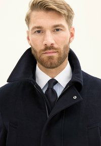 Laivastonsininen villakangas takki korkealla kauluksella, joka sisältää vetoketjukiinnityksen ja sivutaskun. Yllä valkoinen kauluspaita ja tekstuurinen solmio.