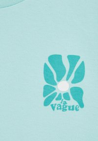 Světle modré bavlněné tričko s velkým stylizovaným grafikou teal květu a textem "la vague" ve stejné barvě, s bílým středem.