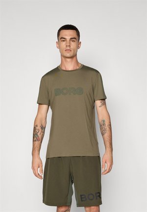 Mand med kort hår og tatoveringer iført olivengrønt BORGA mærket t-shirt og shorts foran en ensfarvet grå baggrund.
