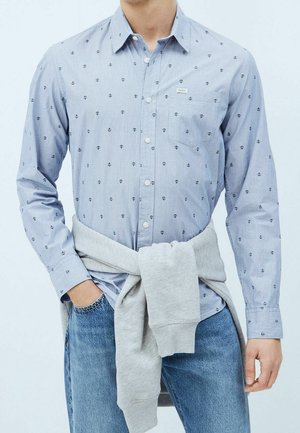 Man draagt lichtblauw overhemd met knopen en ankerpatronen, blauwe spijkerbroek en een grijze trui om de taille gebonden, hand in de zak.