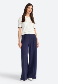 Polo en tricot blanc à manches courtes avec un motif texturé, associé à un pantalon large navy avec des rayures blanches sur les côtés.