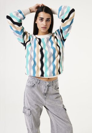 Mehrfarbiger, kurz geschnittener Pullover mit Wellenmuster in Türkis, Beige, Grau und Schwarz; kombiniert mit hellgrauen Cargo-Hosen mit Seitentaschen.