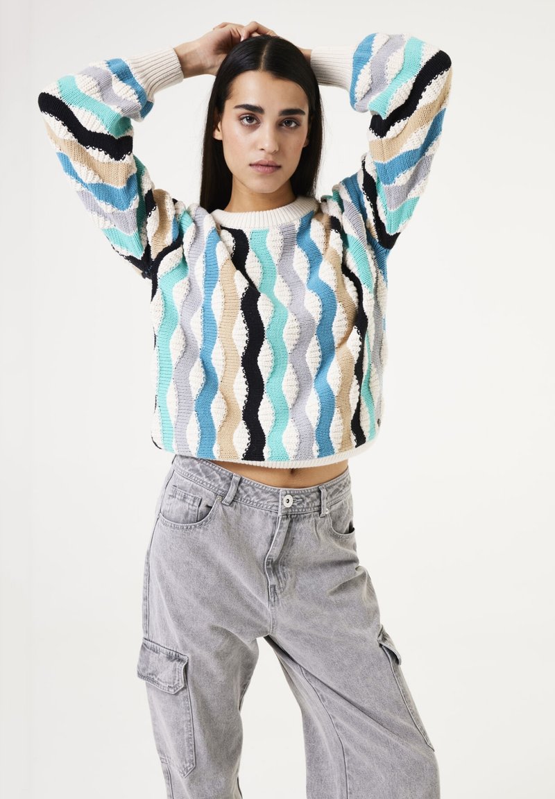 Multikleurige cropped sweater met golvenpatroon in teal, beige, grijs en zwart; gecombineerd met lichtgrijze cargobroek met zijzakken.