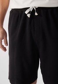 Schwarze Shorts mit Waffelstruktur, einem elastischen Bund und weißen Kordelzügen. Enthält Seitentaschen und ein lässiges Design.