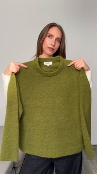 Olivově zelený svetr s texturovaným pleteným povrchem, s vysokým kulatým výstřihem a dlouhými rukávy, držený modelem proti světlému pozadí.