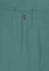 Green fabric with a small round silver button stitched near the top center, showing detailed texture and seam lines.