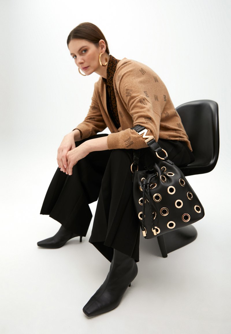 Černá taška ve tvaru kbelíku s pozlacenými oky, hnědý cardigan s textovým vzorem, černé široké kalhoty a černé špičaté lodičky na podpatku.