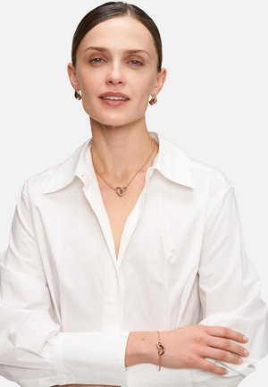 Schmuck aus Roségold umfasst eine herzförmige Anhänger-Halskette und ein Armband mit einem verbundenen Charm, die über einem weißen Hemd mit Knöpfen getragen werden.