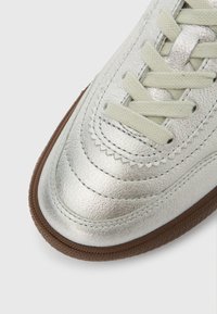 Sneaker metallico argento con lacci beige e suola in gomma marrone, con cuciture dettagliate lungo l'area delle dita, su sfondo bianco.