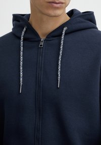 Donkerblauwe zip-up hoodie met een capuchon, patroonde trekkoorden en een rits met metalen accent. Zachte textuur en minimaal ontwerp.