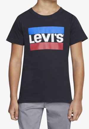 Dziecko w czarnej koszulce Levi's z niebiesko-czerwonym prostokątnym logo i jasnoszarych spodniach, stoi z rękami swobodnie opuszczonymi wzdłuż ciała.