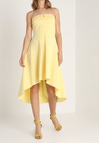 Vestido amarillo sin tirantes con un dobladillo asimétrico y falda plisada. Hecho de una tela suave. Con sandalias amarillas a juego.