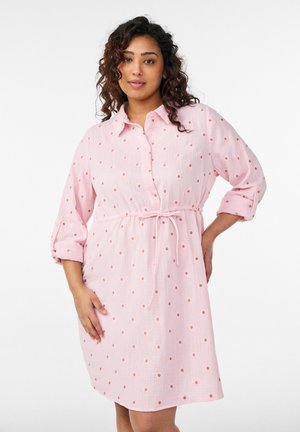 Žena s kučeravými vlasmi v svetloružových dlhých rukávoch šatách s malými vyšívanými bodkami, stojaca s jednou rukou na boku.